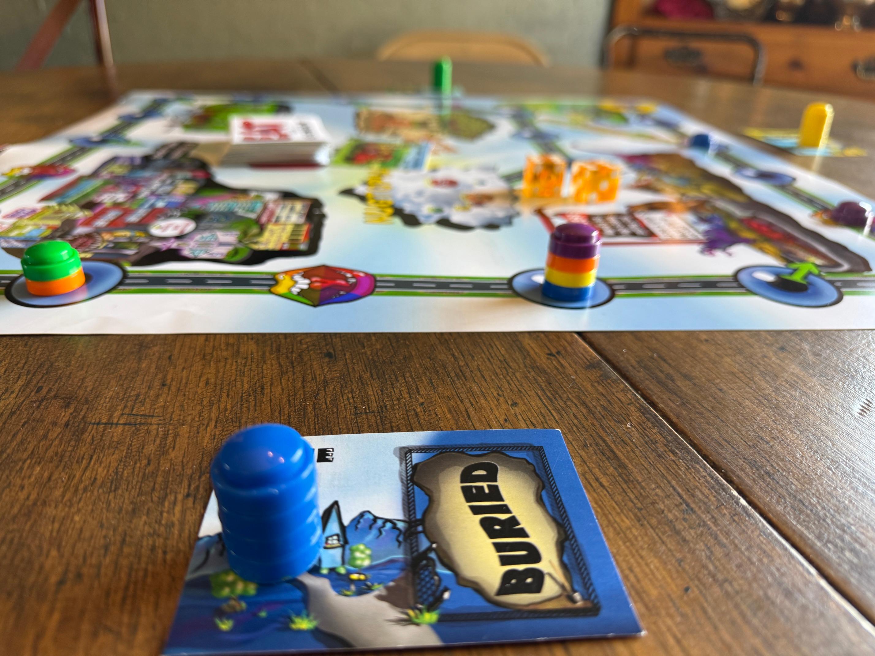 The Eat Your Neighbor board mid-game on a wooden table, with colorful player pawns, dice, event cards, and a Home card marked Buried in the foreground